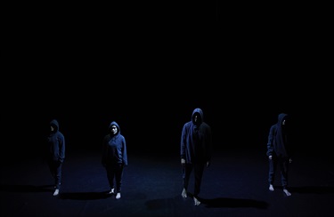 Four people wear dark clothing from head to toe and stand across a darkly lit black stage.